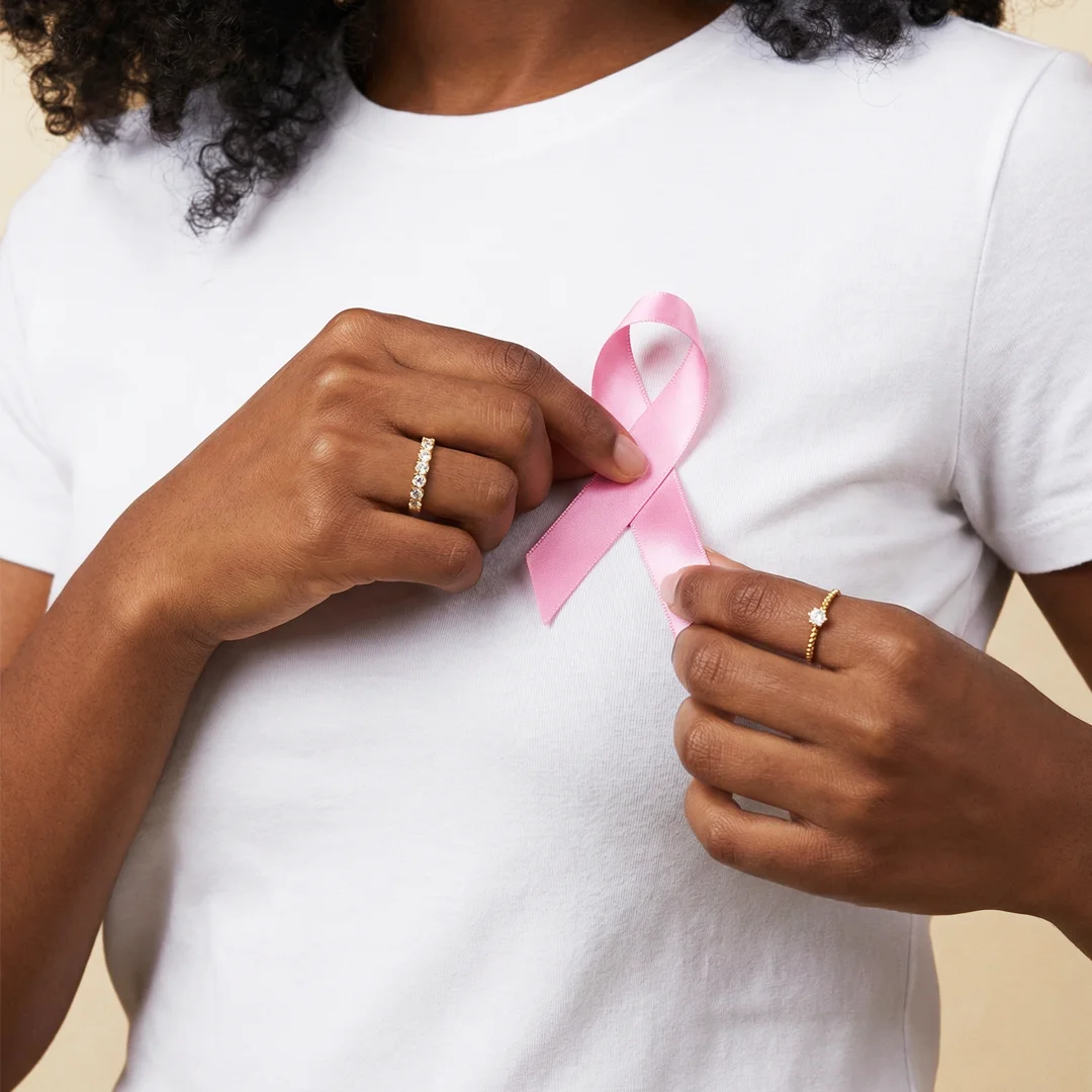 Frau im weißen Shirt hält eine rosa Schleife als Symbol gegen Brustkrebs, an ihren Händen zarte goldene Ringe von Hope22 – Hope22 spendet für Krebsprojekte