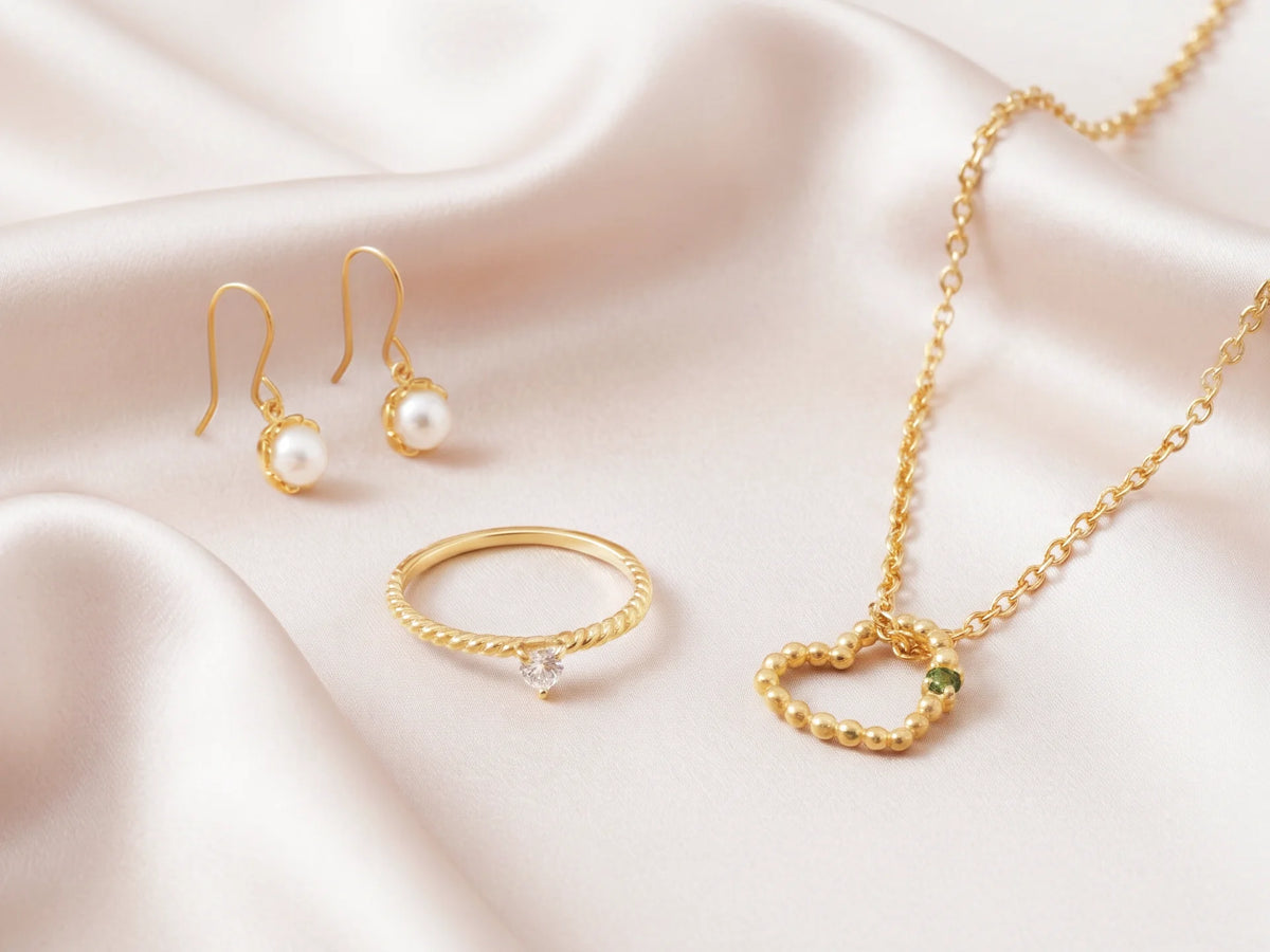 Goldener Schmuck von Hope22 auf cremefarbener Seide – Perlenohrringe, Ring mit Stein und Kette mit Herzanhänger